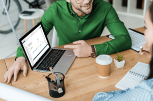 Profissional de tecnologia de blusa verde aponta para um notebook em cima da mesa apresentando as vantagens da solução Multicloud Mendix